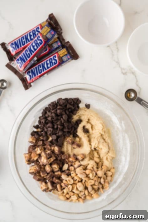 Chocolate chips, peanuts, and Snickers being added to cookie dough.