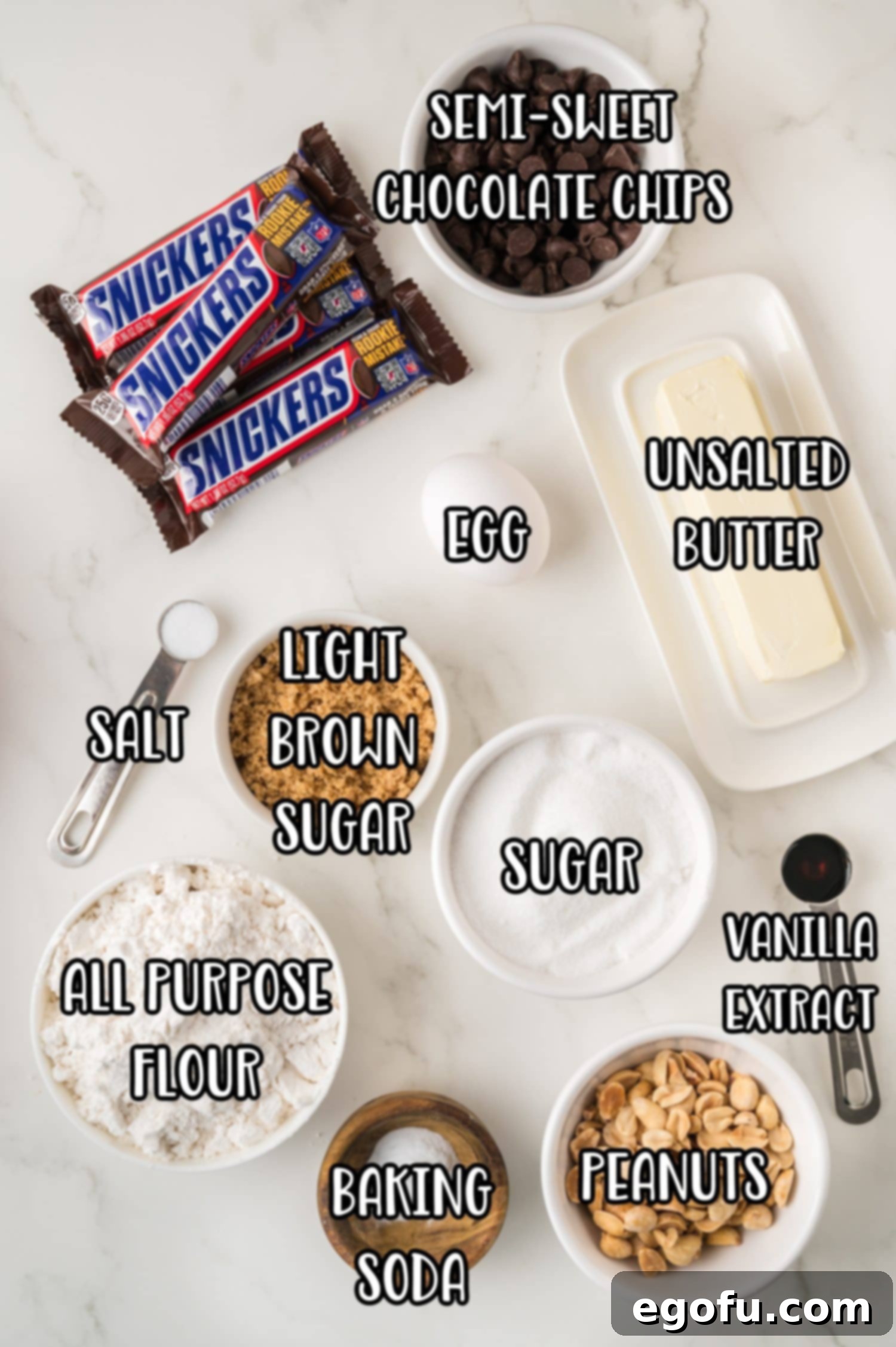 A flat lay of all the essential ingredients for Snickers Cookies: all-purpose flour, semi-sweet chocolate chips, unsalted butter, salt, granulated sugar, light brown sugar, an egg, vanilla extract, baking soda, chopped peanuts, and Snickers bars.