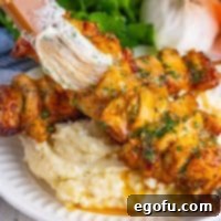 A close-up image of a brush applying sauce to glistening chicken skewers served over a bed of mashed potatoes, highlighting the rich garlic parmesan coating.