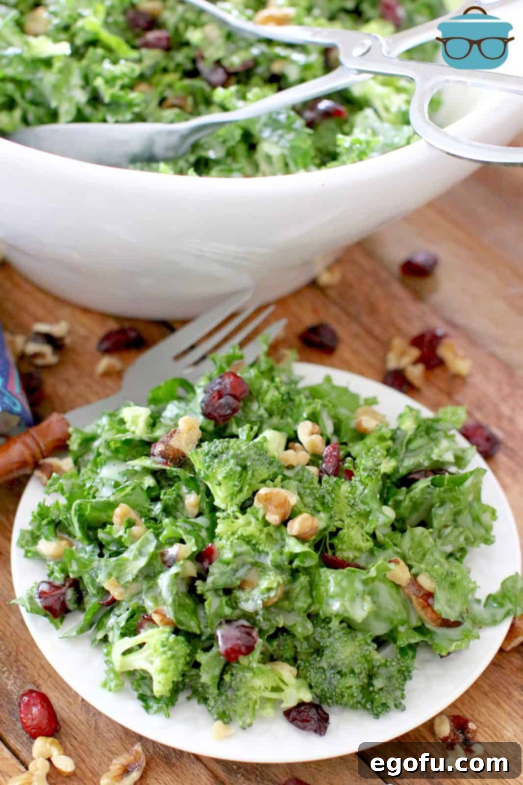 A vibrant kale and broccoli salad, garnished with walnuts and dried cherries, served on a small round white plate. A white bowl sits in the background on a rustic wood surface.