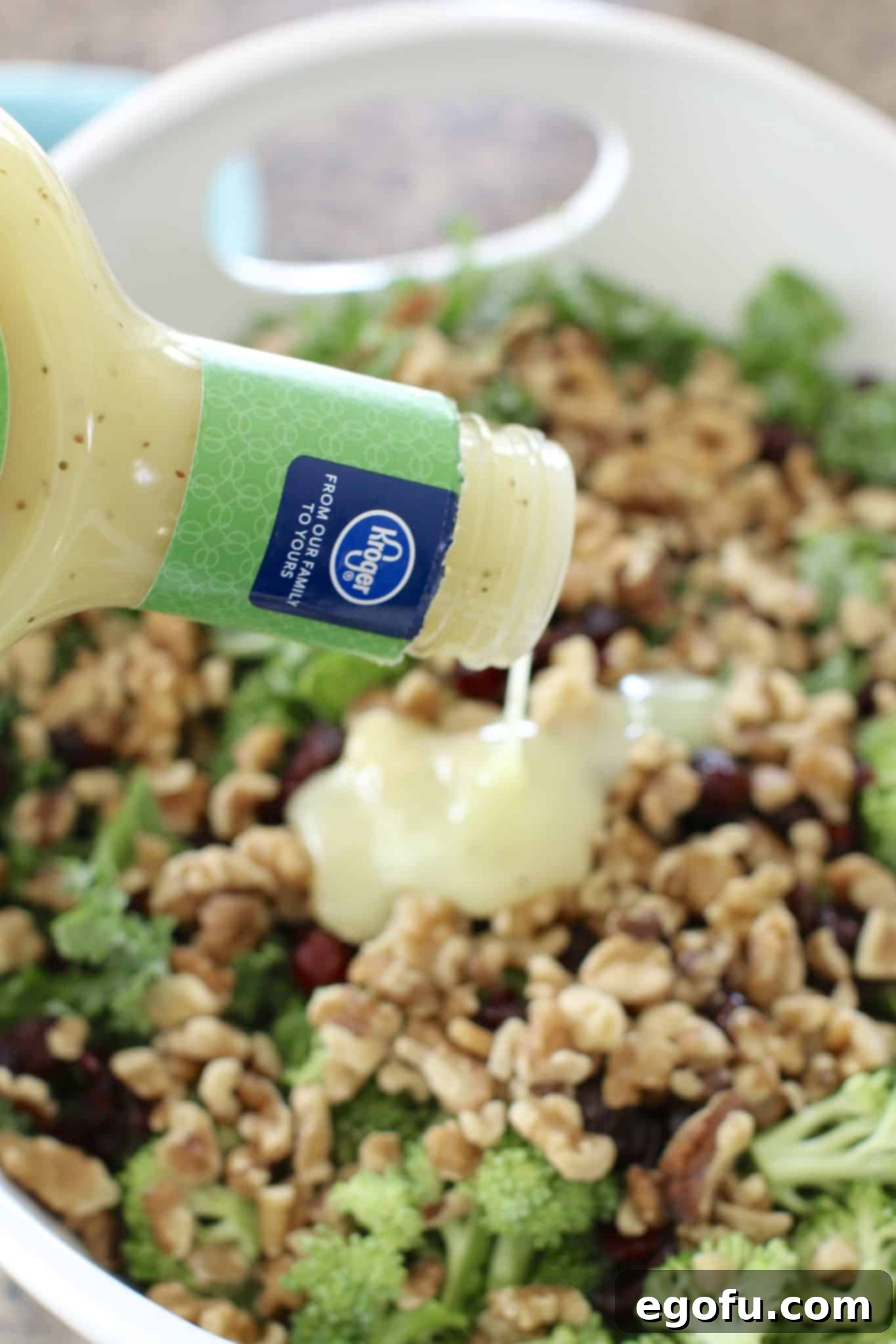 Sweet and sour dressing being poured generously over the mixed kale, broccoli, walnuts, and cranberries in a large salad bowl.