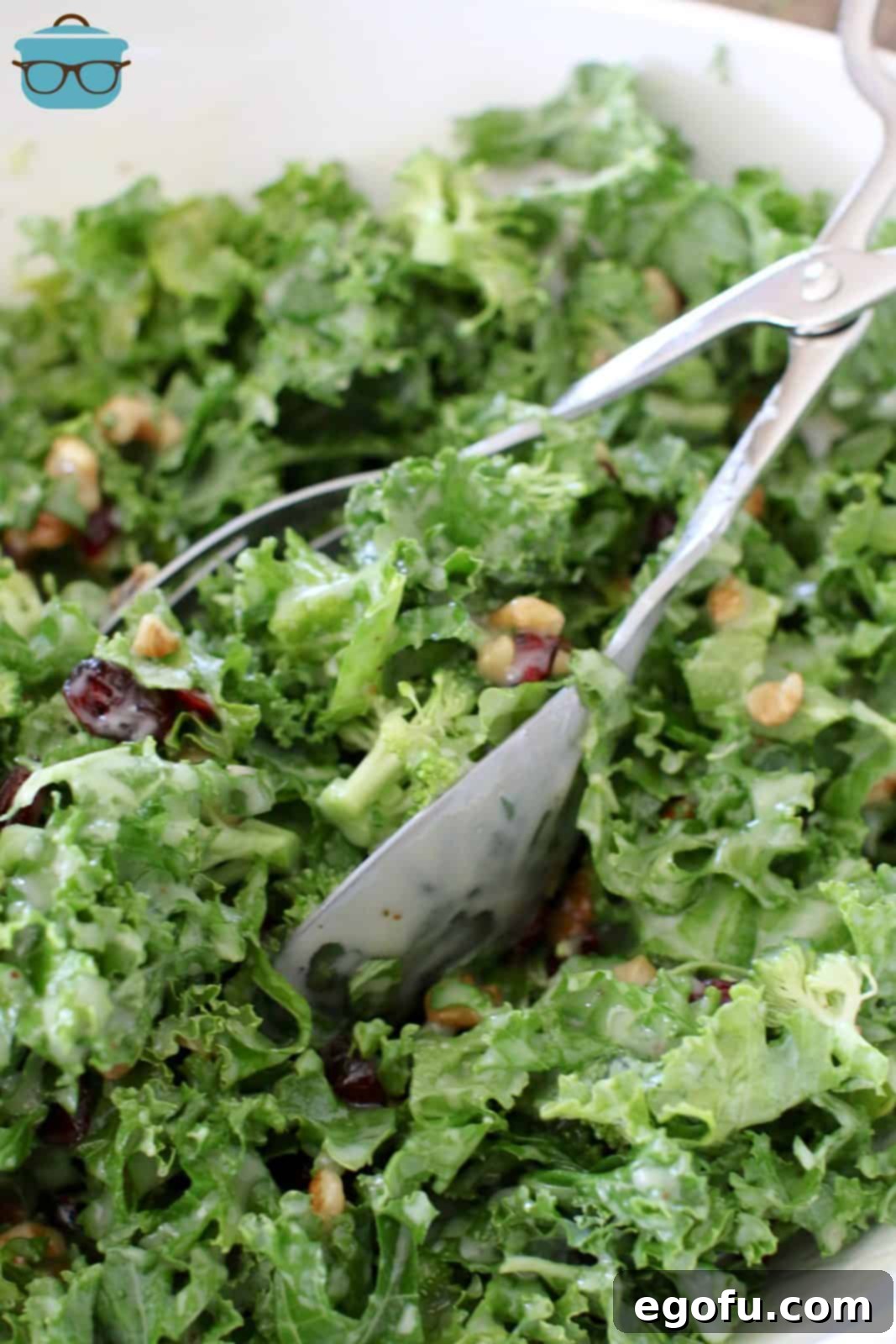 A pair of silver salad tongs resting in a large white bowl brimming with freshly prepared kale salad, indicating it's ready to be served.