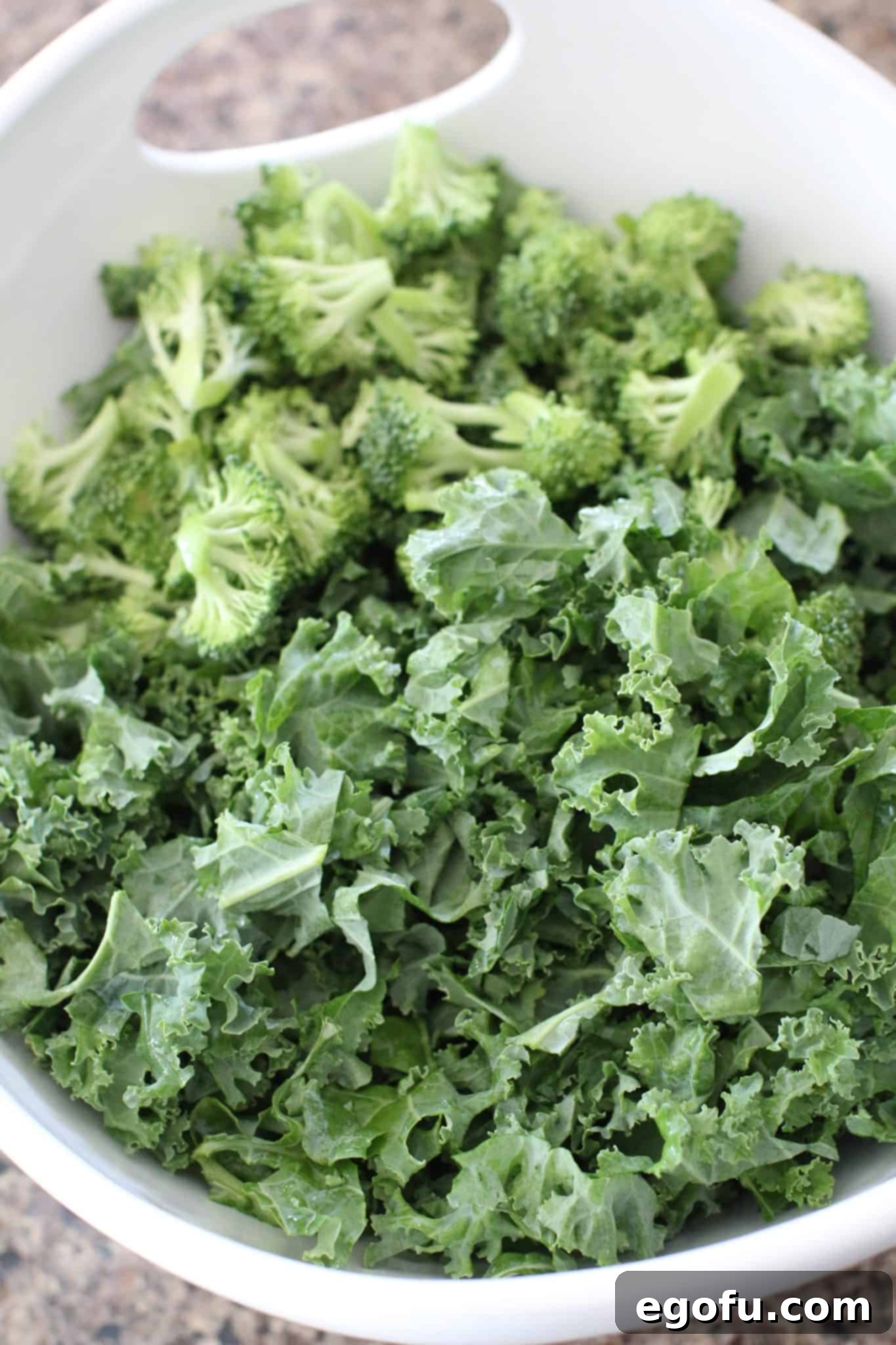 A large white bowl with handles containing a mix of vibrant kale and green broccoli florets, creating the salad's base.