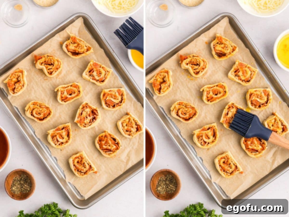 Unbaked pinwheel slices are arranged on a parchment-lined baking sheet. A pastry brush applies egg wash, and then grated Parmesan cheese is sprinkled over them.