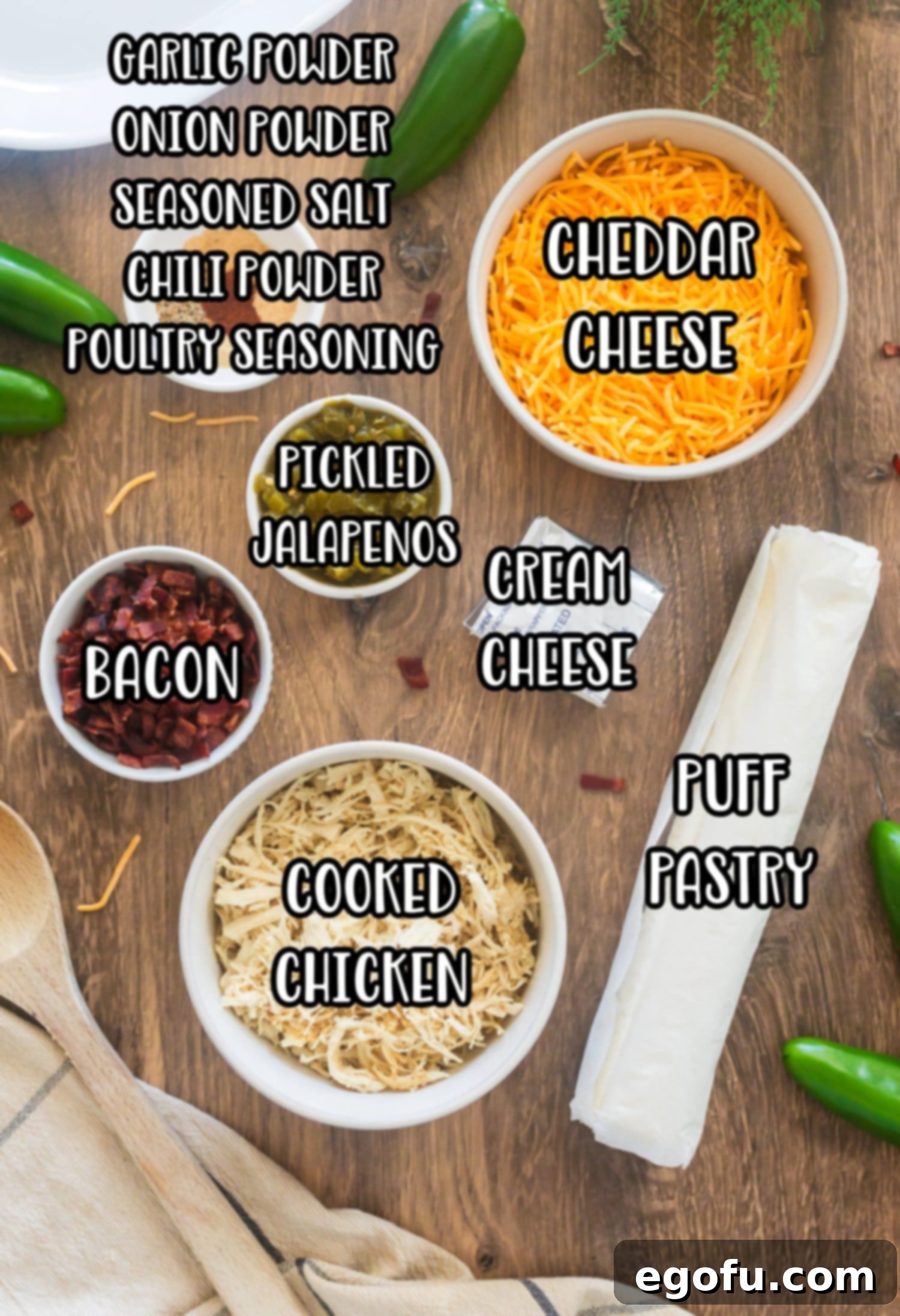 A flat lay photograph showcasing the ingredients for Baked Jalapeño Popper Pinwheels: puff pastry, diced pickled jalapenos, shredded cheddar cheese, cream cheese, cooked shredded chicken, chopped bacon, and various spices.