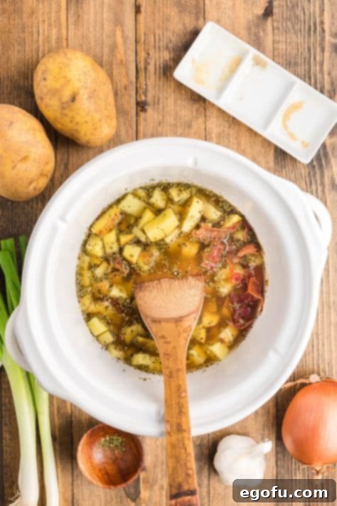 A wooden spoon gently stirring the contents of a small slow cooker filled with simmering potato soup, indicating the cooking process.