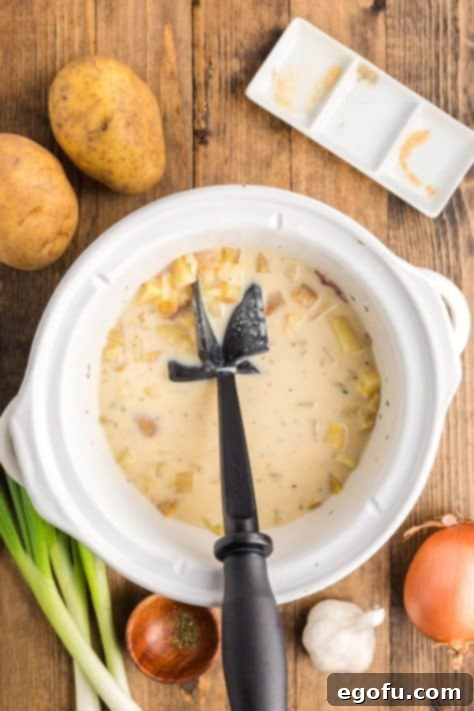 A potato masher in action, mashing tender potatoes within the creamy soup in a slow cooker to achieve desired texture and thickness.