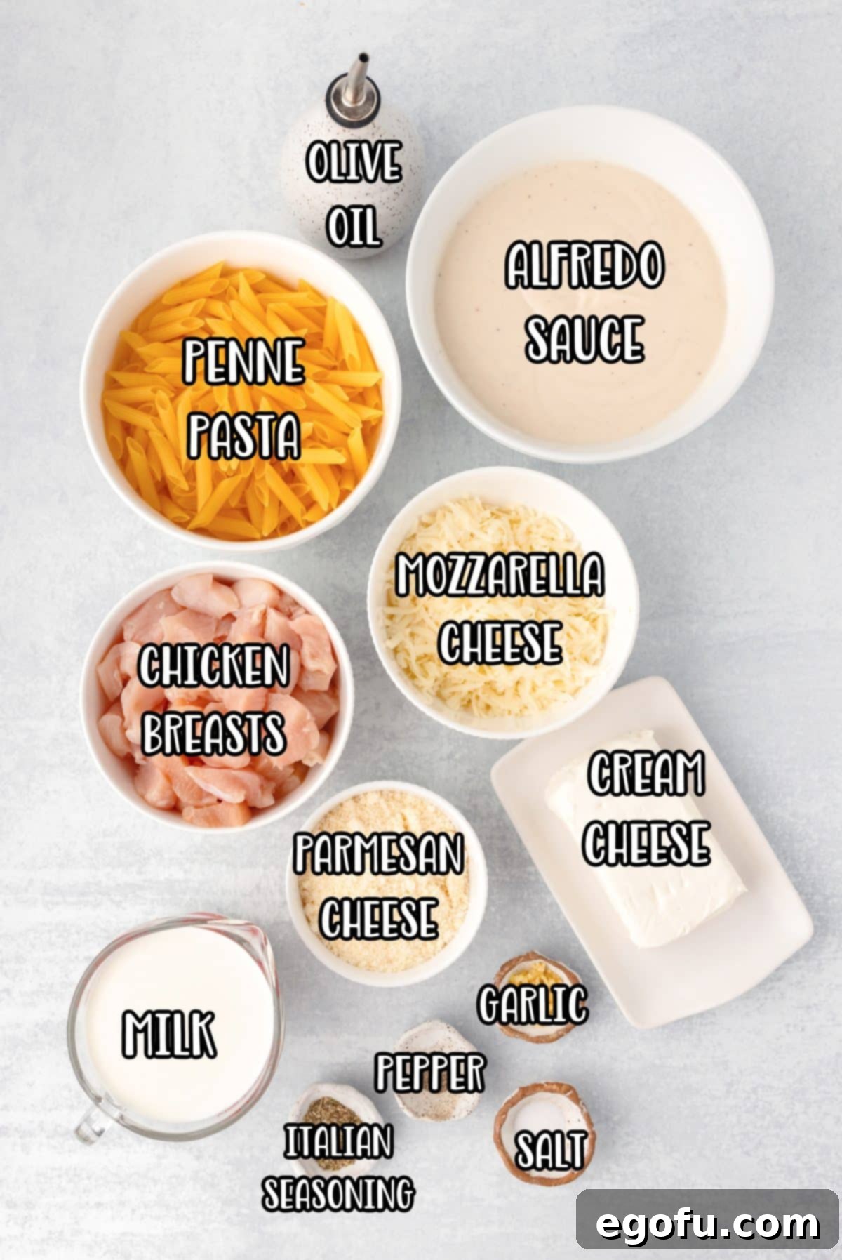 Penne pasta, Alfredo sauce, mozzarella cheese, chicken breast pieces, parmesan cheese, cream cheese, milk, garlic powder, salt, pepper, olive oil, minced garlic, Italian seasoning, and parsley are laid out as ingredients for Chicken Alfredo Casserole.