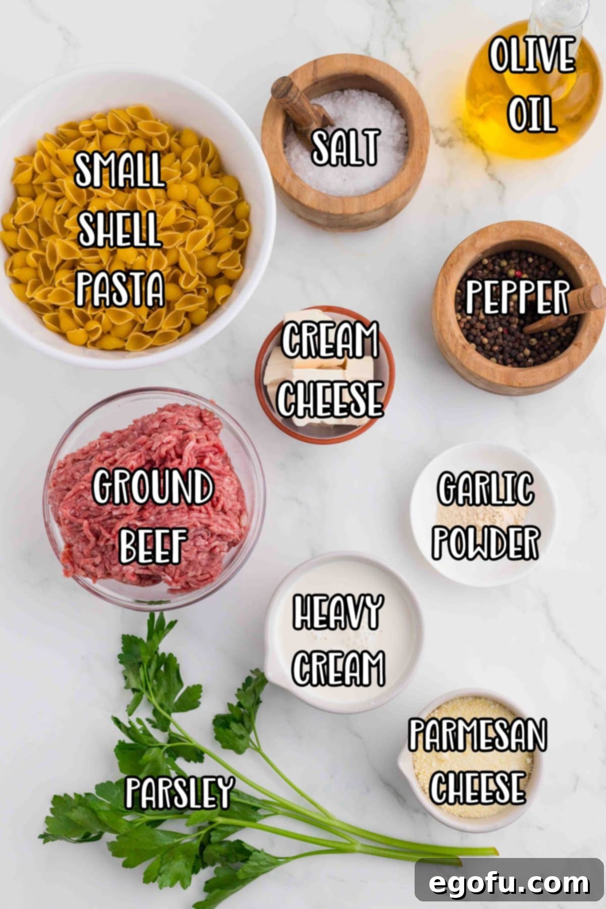 A flat lay photograph showcasing all the ingredients for Ground Beef Alfredo: small shell pasta, ground beef, heavy cream, cream cheese, parmesan cheese, olive oil, garlic powder, salt, pepper, and fresh parsley.