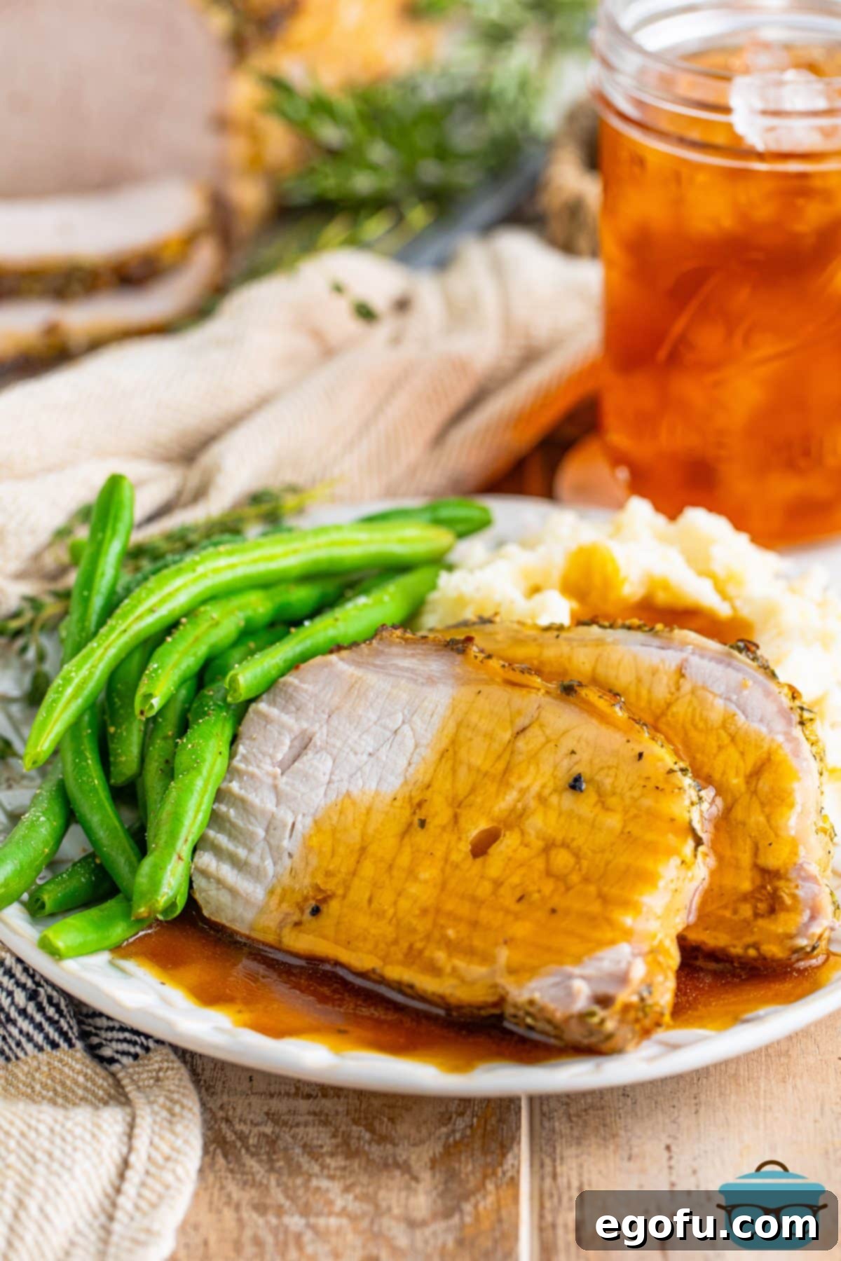 A few slices of gravy-coated herb crusted pork roast on a plate with creamy mashed potatoes and vibrant green beans.