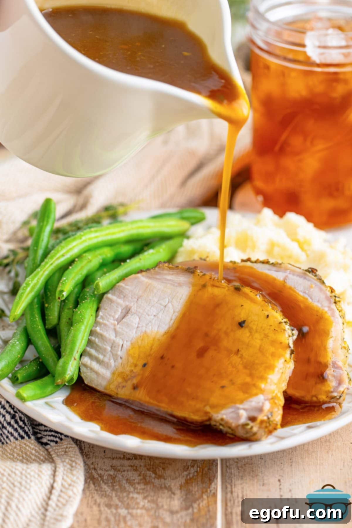 Rich gravy being poured over a few slices of Herb Crusted Pork Roast on a plate, accompanied by green beans.