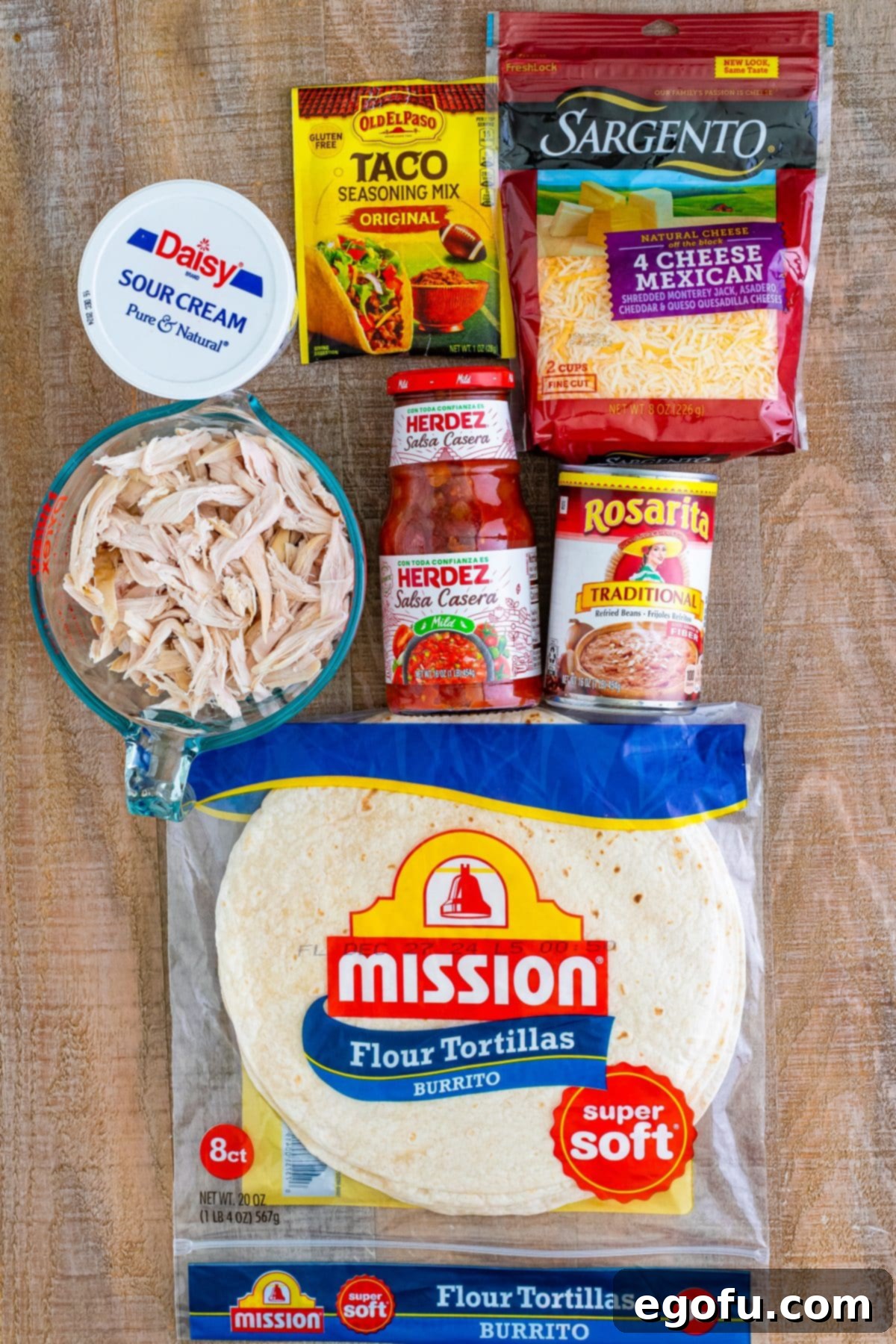A vibrant flat lay of all the essential ingredients for making Chicken Chimichangas: large burrito tortillas, a whole rotisserie chicken, sour cream, taco seasoning, shredded Mexican cheese, refried beans, savory salsa, and a bottle of cooking oil, neatly arranged on a wooden surface.