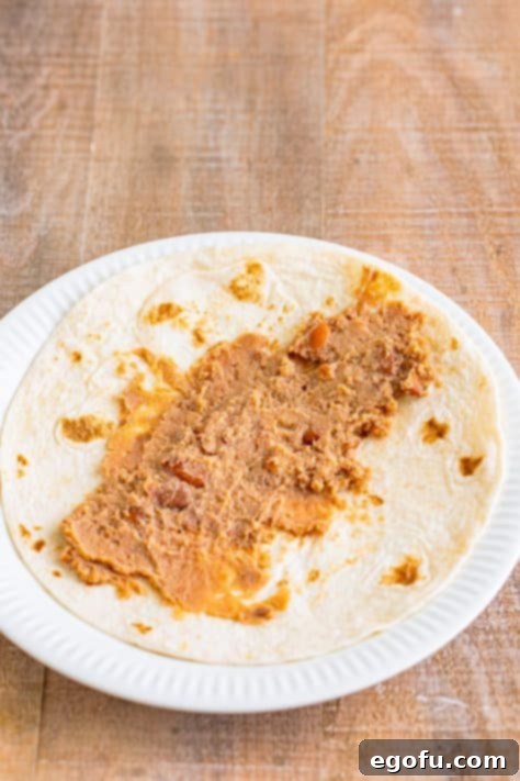 Creamy refried beans spread neatly down the center of a tortilla on a plate.
