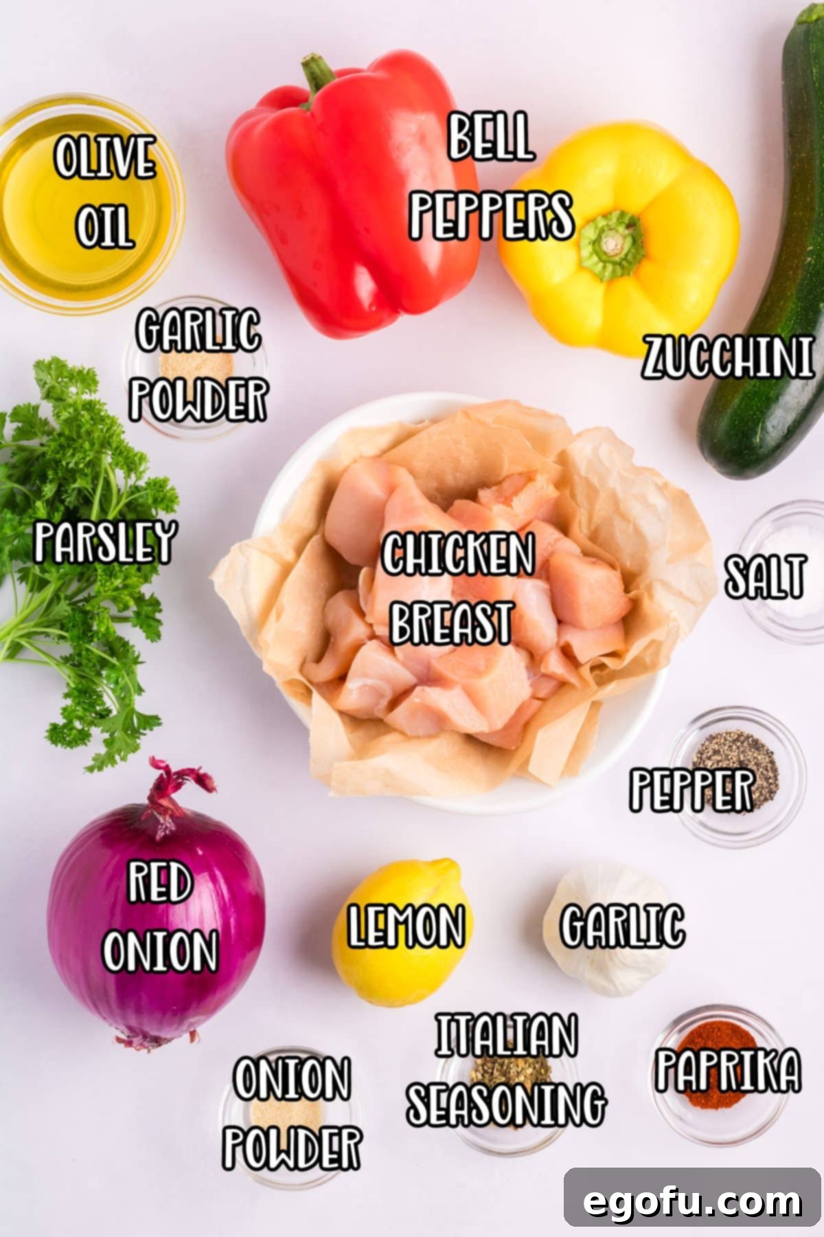 A beautifully arranged selection of fresh ingredients including red peppers, yellow peppers, cubed chicken breast, olive oil, sliced zucchini, red onion, minced garlic cloves, lemon juice, paprika, Italian seasoning, salt, pepper, onion powder, garlic powder, and fresh parsley, laid out for Baked Chicken Skewers preparation.