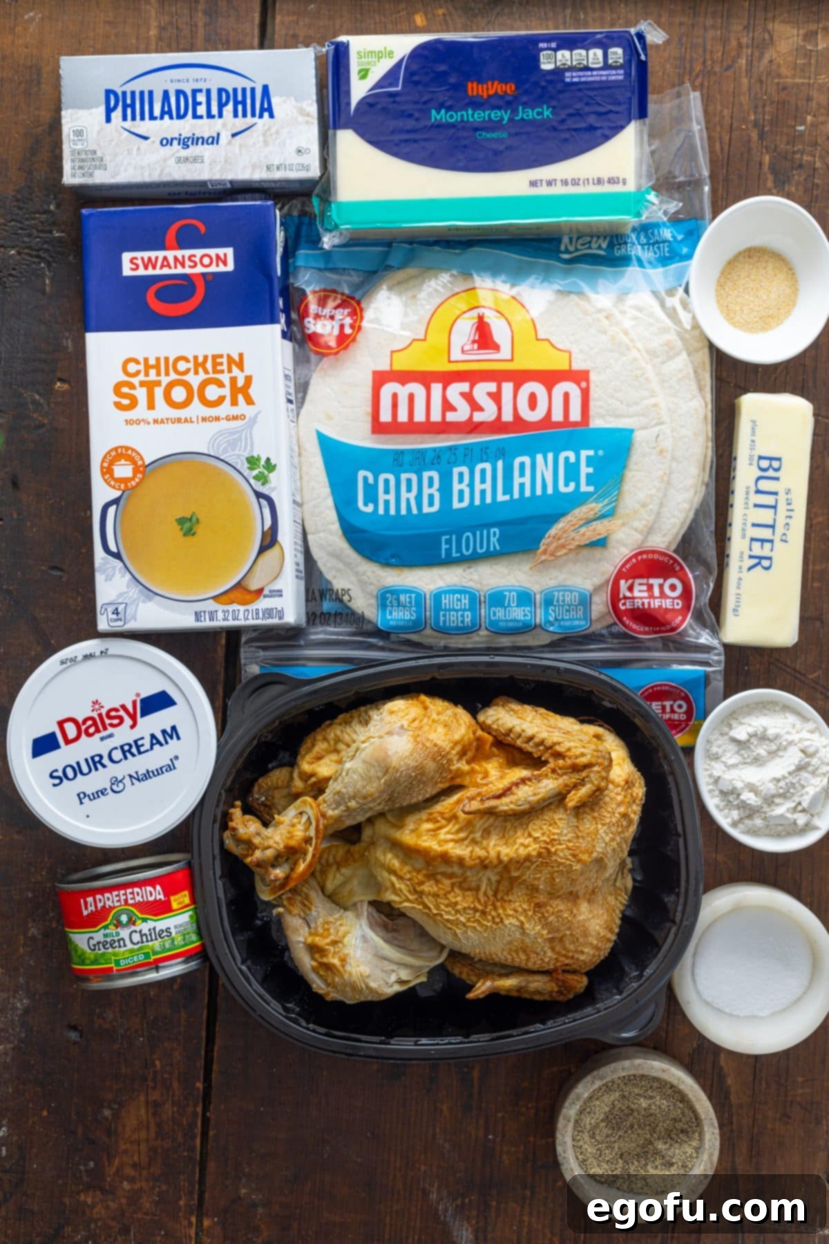 A flat lay of all the fresh ingredients needed for White Chicken Enchilada Casserole, including cream cheese, tortillas, cooked chicken, shredded Monterey cheese, garlic powder, salt, ground black pepper, butter, all purpose flour, chicken broth, sour cream, diced green chilies, and fresh parsley.