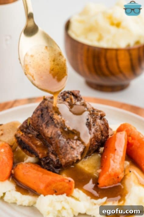 A spoon drizzling gravy over some Crock Pot Short Ribs with veggies on a pile of mashed potatoes.