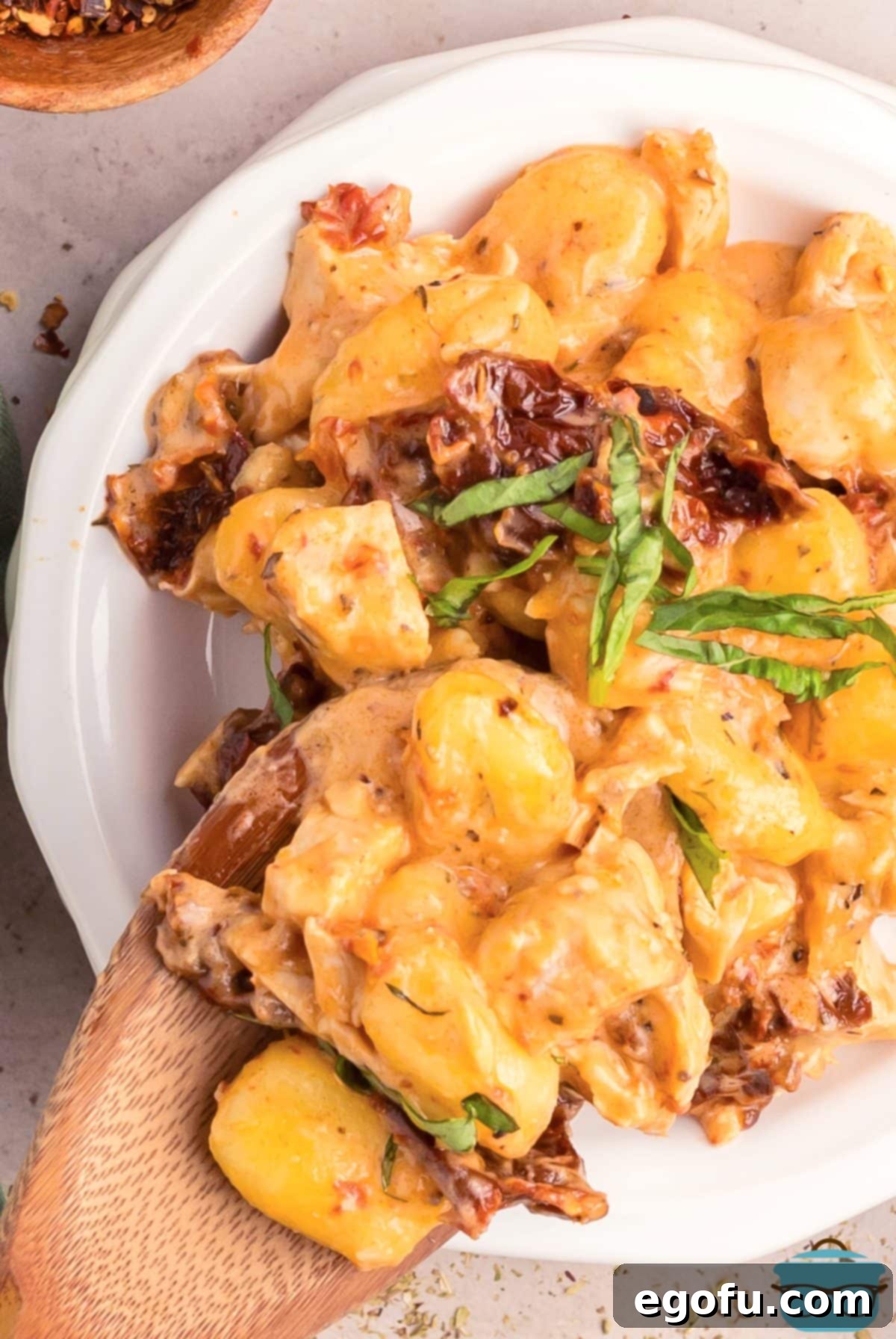 A wooden spoon putting a large serving of Marry Me Chicken Gnocchi on a dinner plate, garnished with basil.