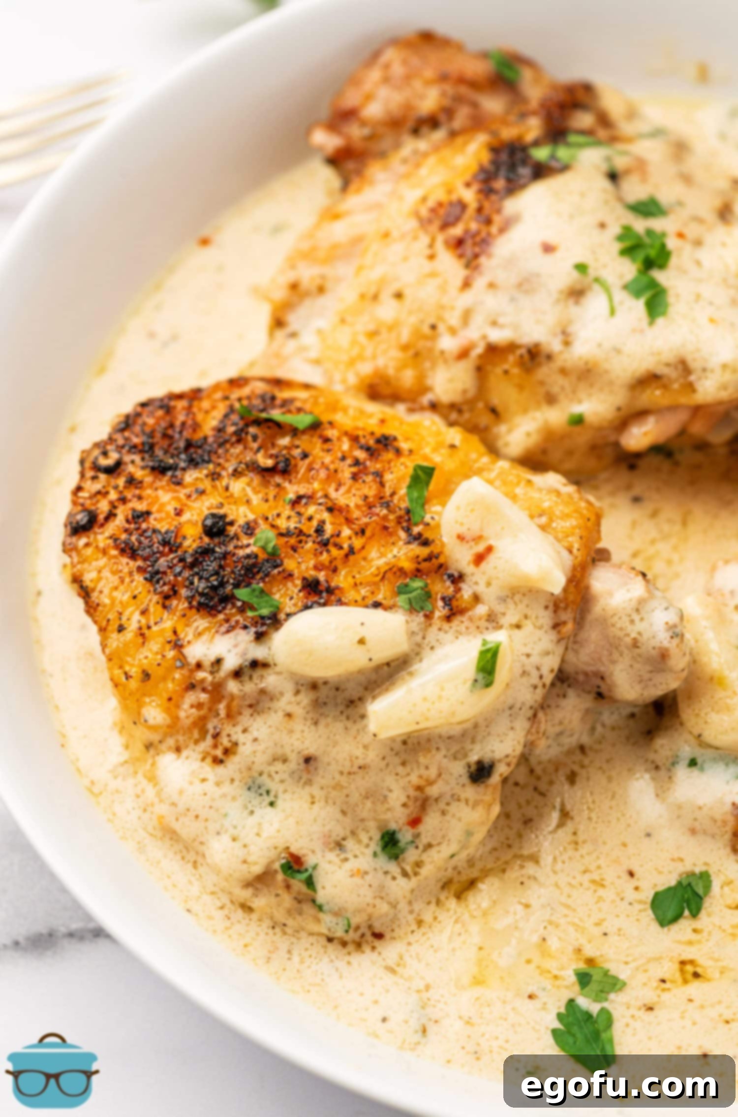 Creamy Garlic Chicken Delight 2 A plate with a golden-brown chicken thigh, generously drizzled with creamy garlic sauce and garnished with fresh garlic slices and herbs.