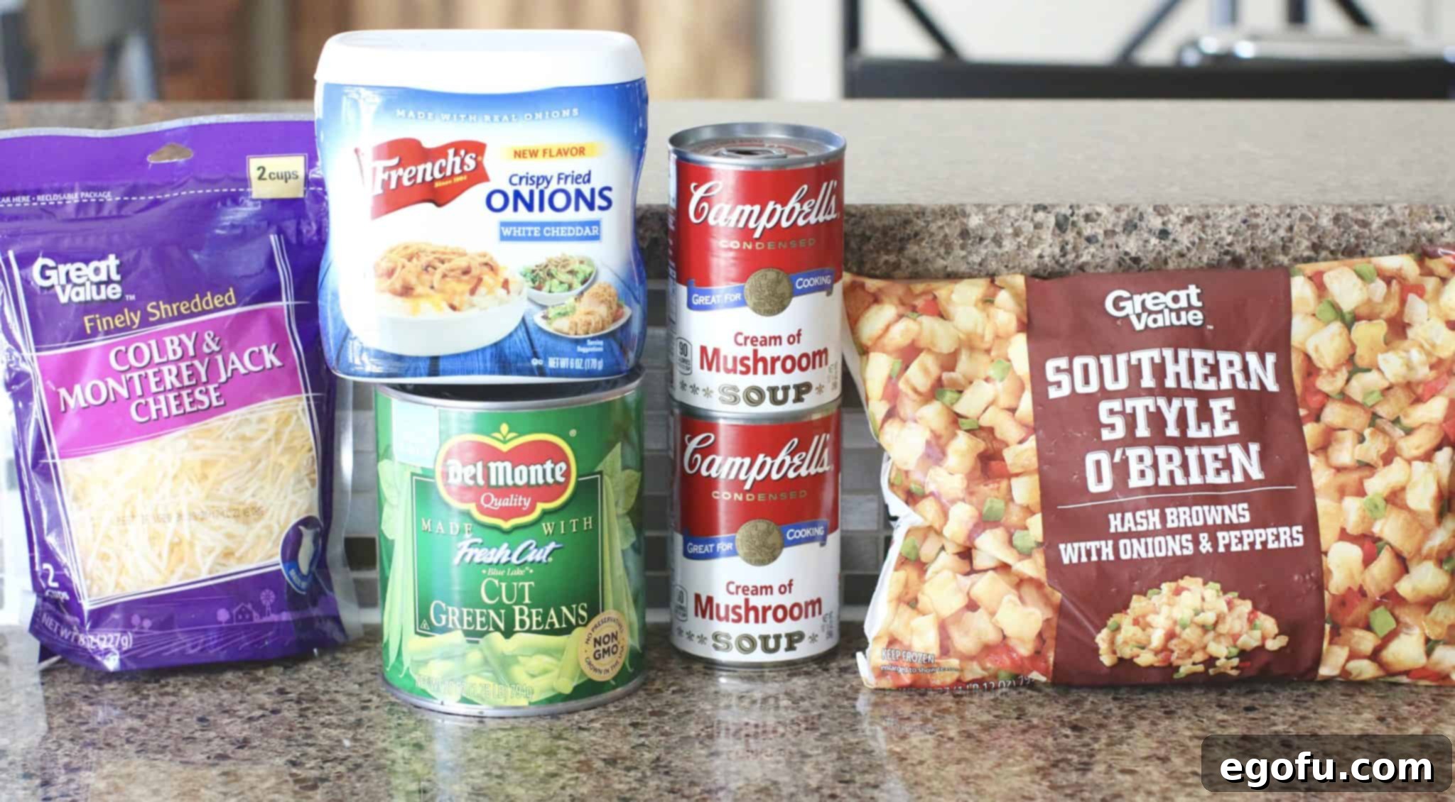 Homestyle Potato Green Bean Bake 4 A vibrant display of key ingredients for Tater Green Bean Casserole, including cans of cream of mushroom soup, a bottle of milk, various seasonings, a bag of frozen O'Brien potatoes, cans of green beans, a container of French's® Crispy Fried Onions, and a bag of shredded cheese, arranged on a rustic wooden surface.