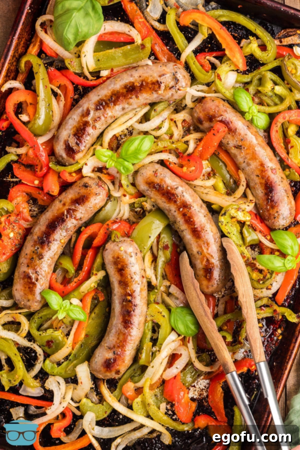 One Pan Sausage and Pepper Bake 6 Tongs grabbing a sausage link from the sheet pan peppers and onions, ready for serving.