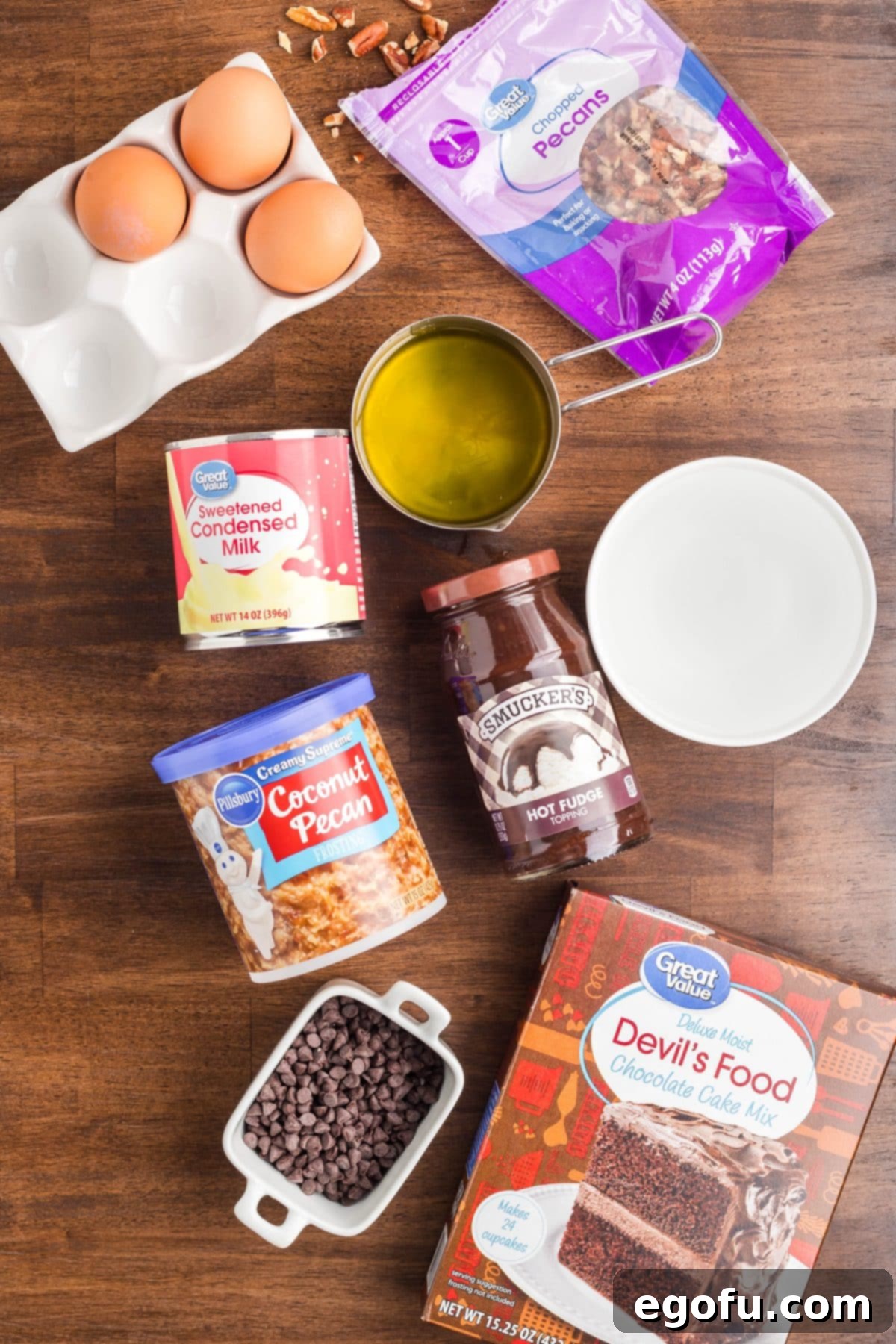 A flat lay photograph displaying all the ingredients needed for German Chocolate Poke Cake: chocolate cake mix, eggs, water, oil, sweetened condensed milk, hot fudge topping, coconut pecan frosting, chopped pecans, and mini chocolate chips.