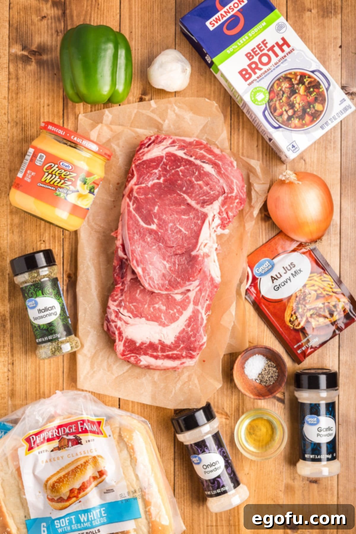 Ribeye steaks, olive oil, salt, pepper, beef broth, au jus gravy, minced garlic, Italian seasoning, garlic powder, sliced onions, green bell peppers, Cheez Wiz, and hoagie rolls.