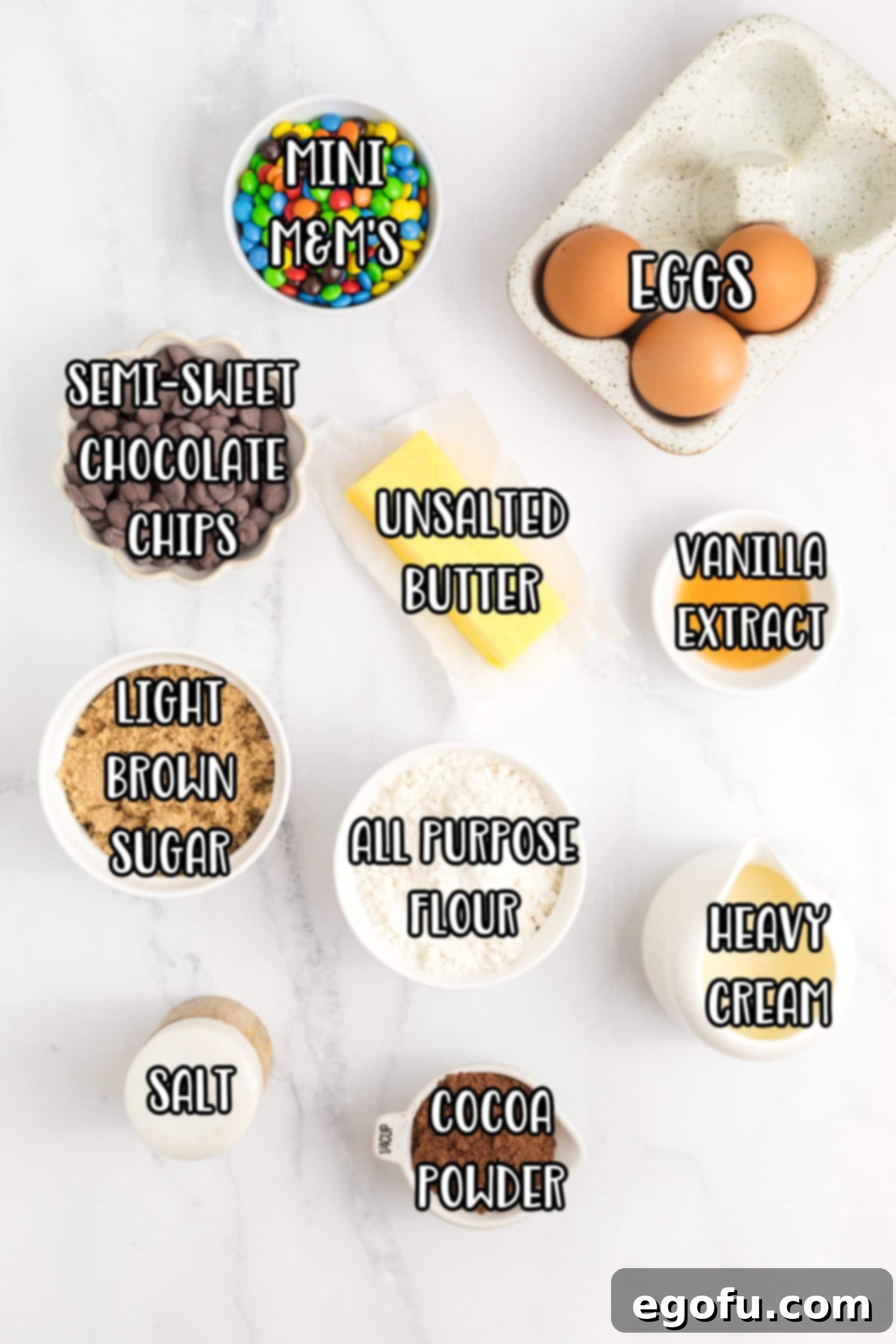 All the ingredients for Cosmic Brownies laid out: eggs, butter, chocolate chips, brown sugar, heavy cream, mini M&M’s, flour, vanilla extract, salt, and cocoa powder.