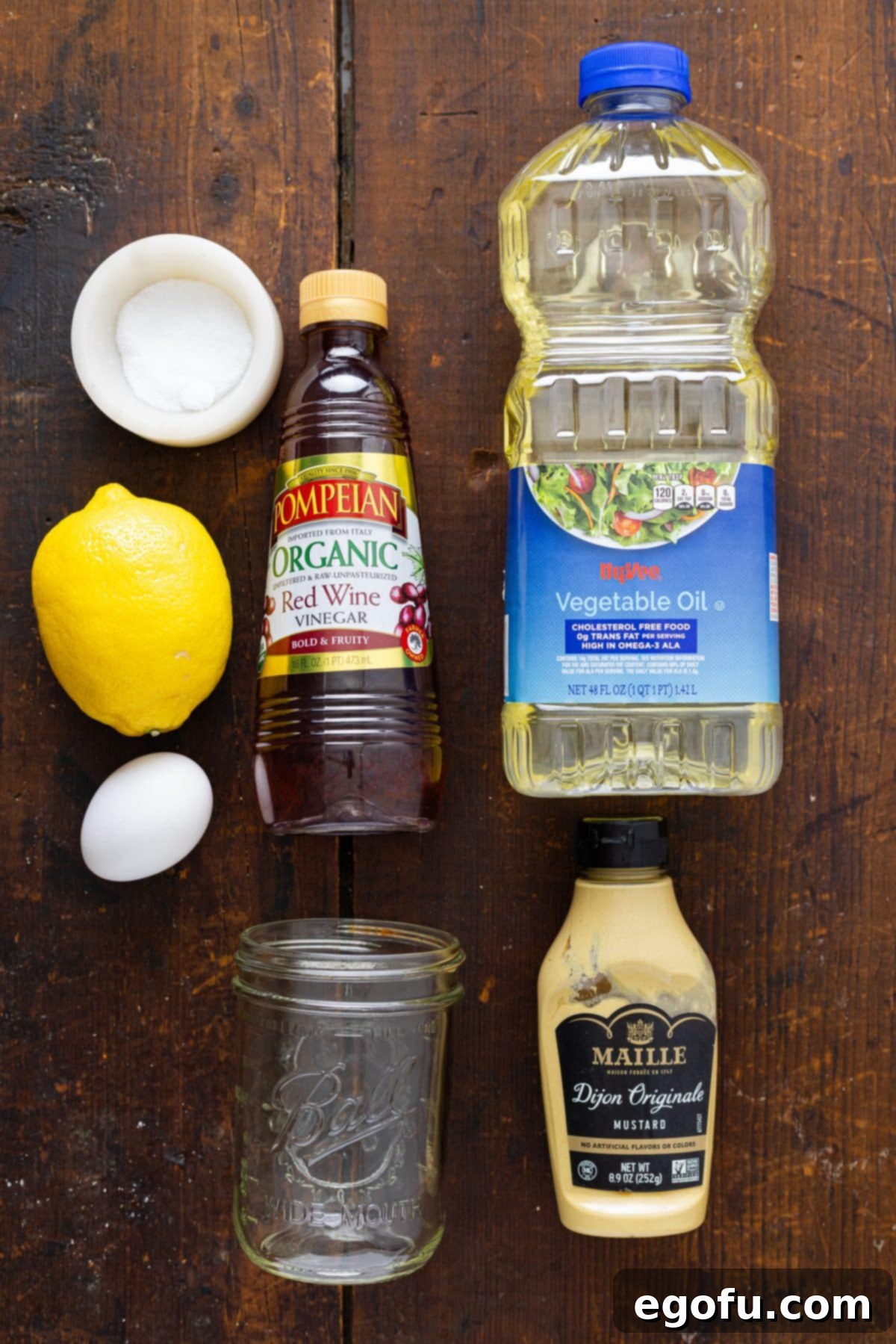 A clean, organized display of all the fresh ingredients required for making Homemade Mayonnaise: a large egg, a bottle of vegetable oil, red wine vinegar, a fresh lemon cut in half, a small dish of salt, a spoonful of Dijon mustard, and an empty mason jar.