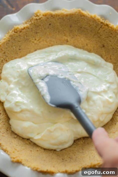 Key lime pie filling added to a golden-brown cookie crust, followed by whipped cream being spread over the top.