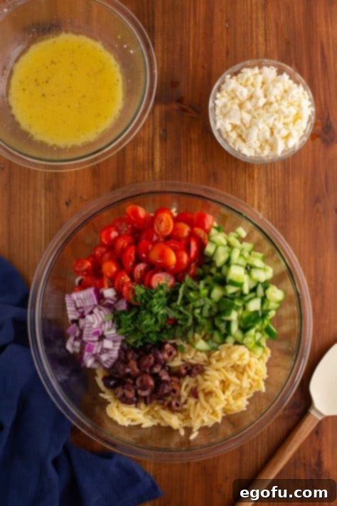 Various fresh ingredients for orzo salad arranged in a large, clear mixing bowl.