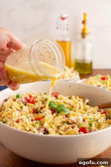 Vinaigrette dressing being drizzled over the mixed orzo salad ingredients in a bowl.