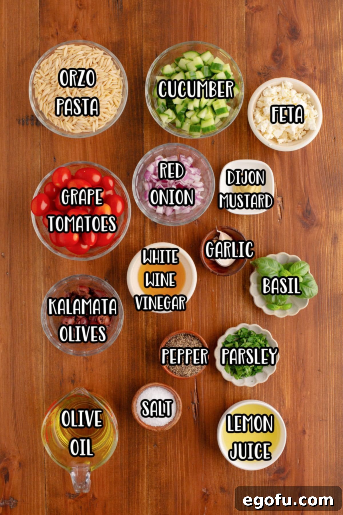 A beautiful flat lay of all the fresh ingredients for Orzo Pasta Salad: orzo, grape tomatoes, basil, parsley, feta cheese, red onion, olive oil, salt, pepper, cucumber, kalamata olives, lemon juice, and Dijon mustard.