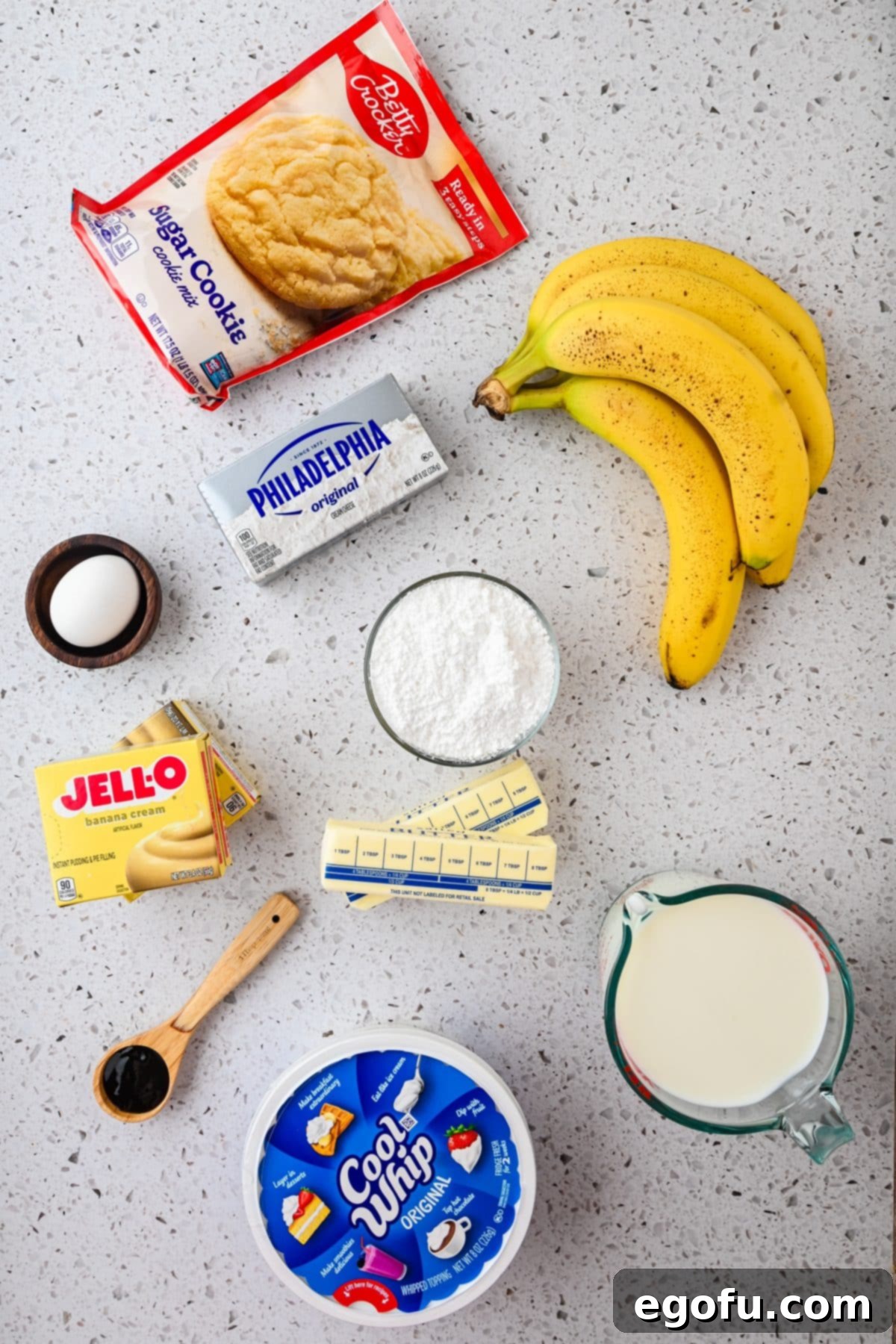 A vibrant flat lay of all ingredients for Banana Pudding Layered Dessert: sugar cookie mix, bananas, Cool Whip, cream cheese, powdered sugar, butter, vanilla extract, egg, milk, and banana pudding mix.