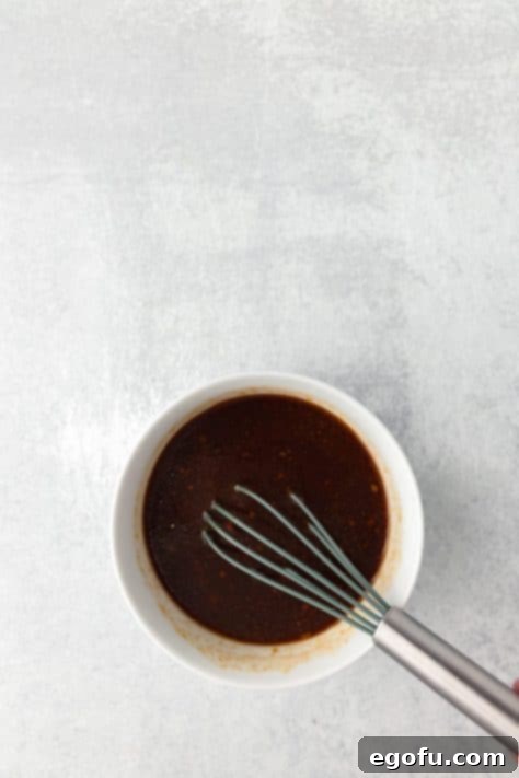 A bowl with pineapple juice, ketchup, honey, soy sauce, apple cider vinegar, sesame oil, garlic, and ginger powder being whisked together.