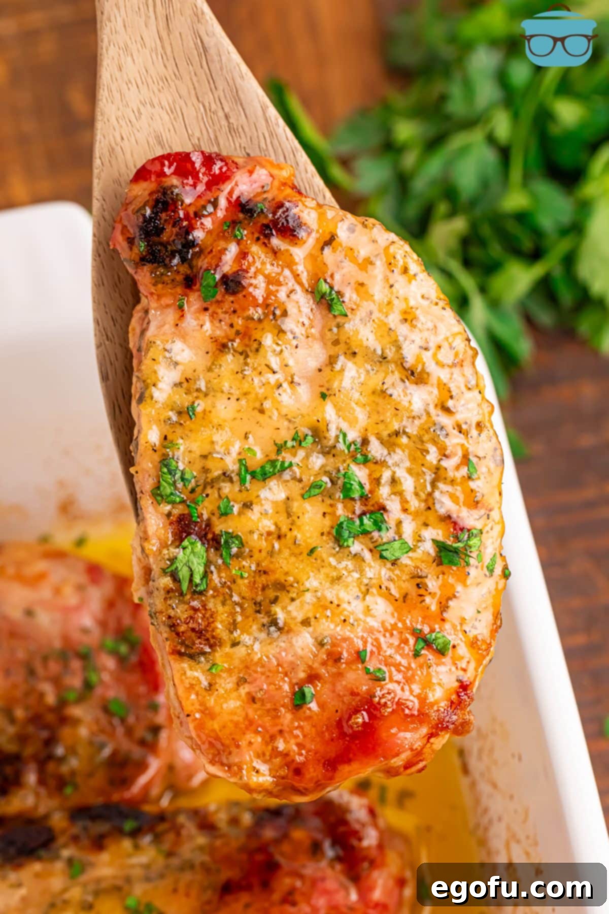 Golden Ranch Butter Pork Chops 2 Wooden serving spoon holding a Ranch Butter Pork Chop above the rest of them in a baking dish.