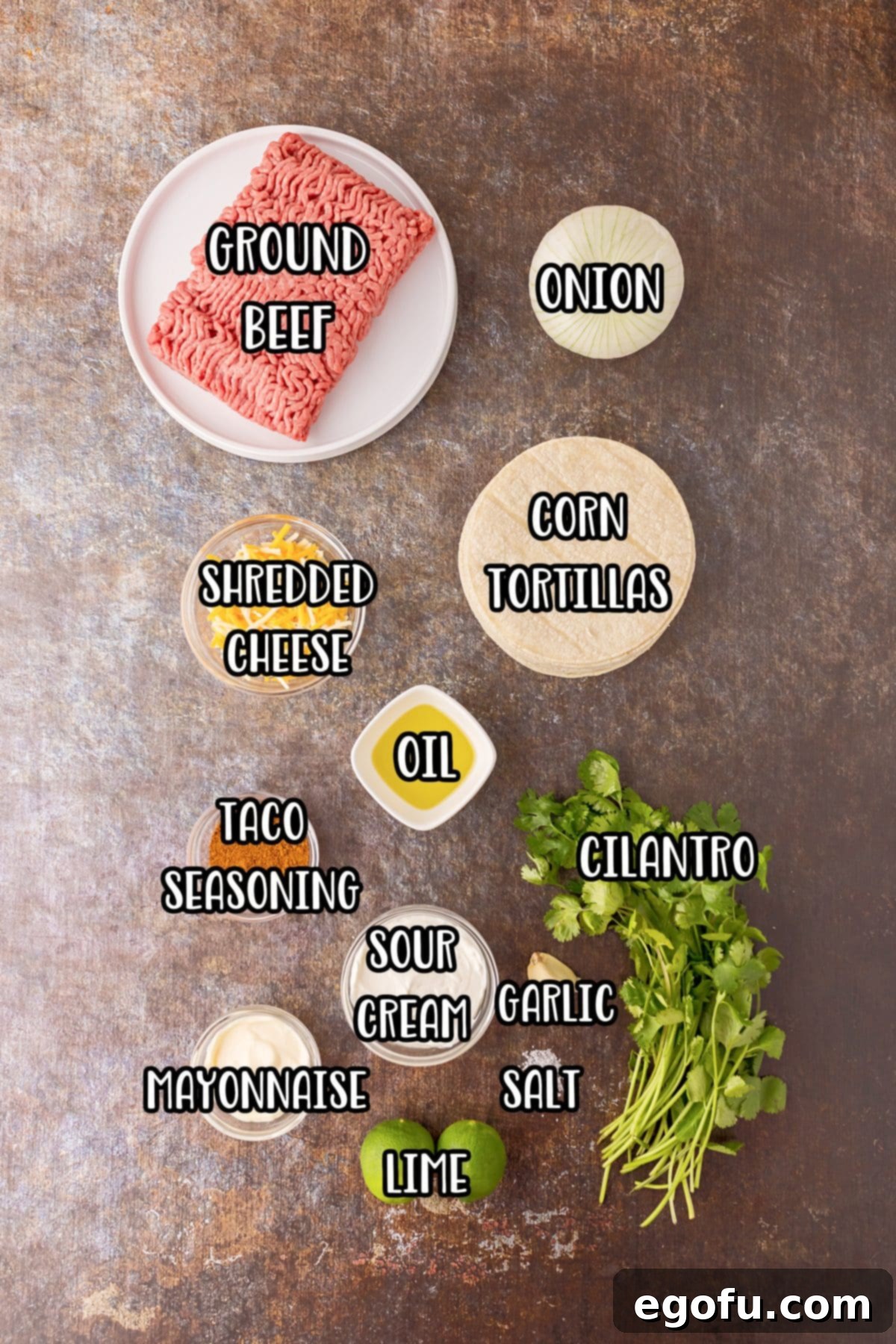 A flat lay of all ingredients for Baked Tacos, including ground beef, onion, corn tortillas, shredded cheese, oil, cilantro, taco seasoning, garlic, salt, sour cream, limes, and mayonnaise.