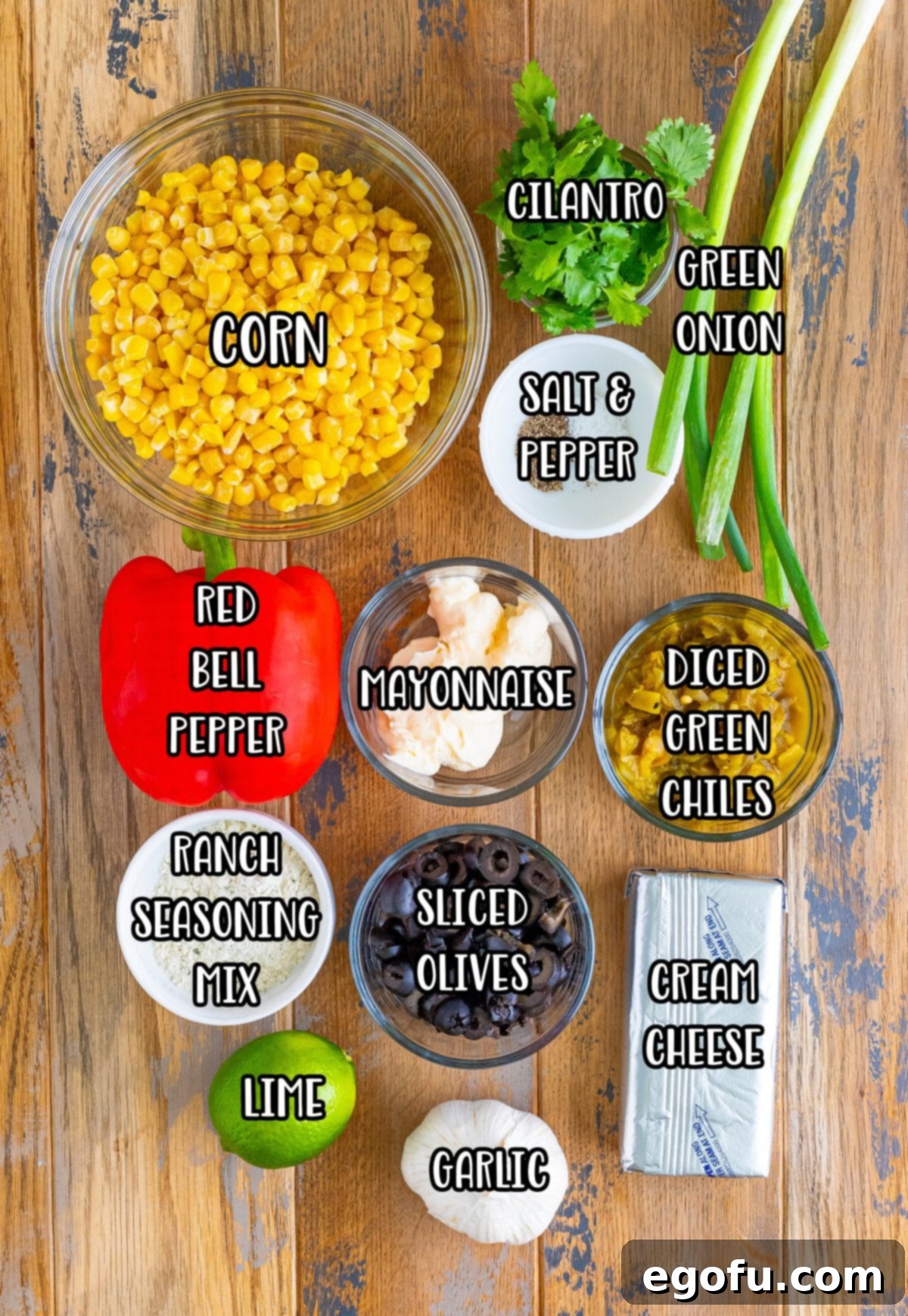 A vibrant flat lay of all fresh and canned ingredients for Poolside Dip, including corn, red pepper, lime, mayonnaise, salt, pepper, green onion, diced green chiles, cilantro, ranch seasoning, black olives, minced garlic, and cream cheese, neatly arranged on a rustic wooden surface.