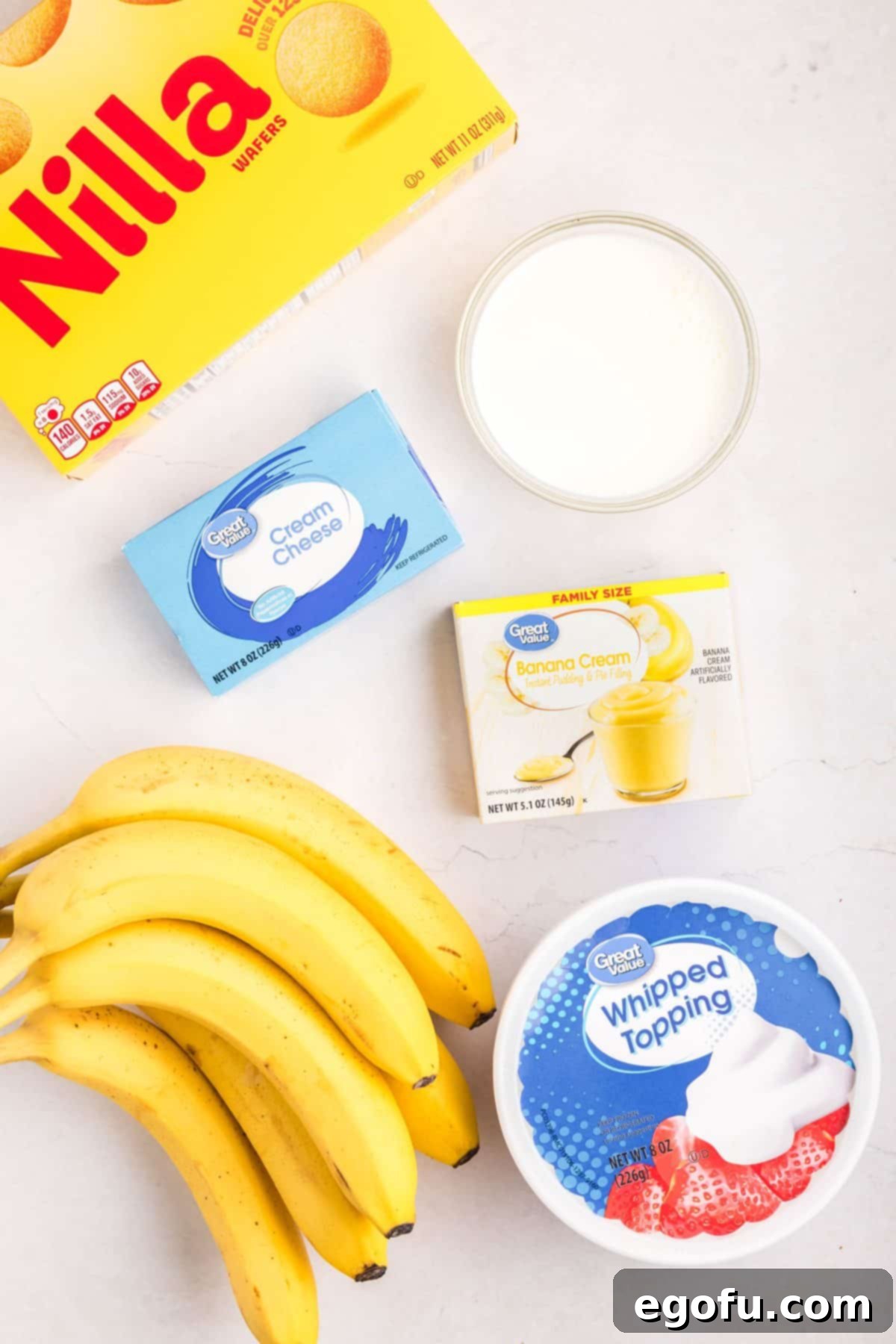 All six key ingredients for No-Bake Banana Pudding laid out: ripe bananas, instant banana pudding mix, Nilla wafers, cream cheese, whipped topping, and whole milk.