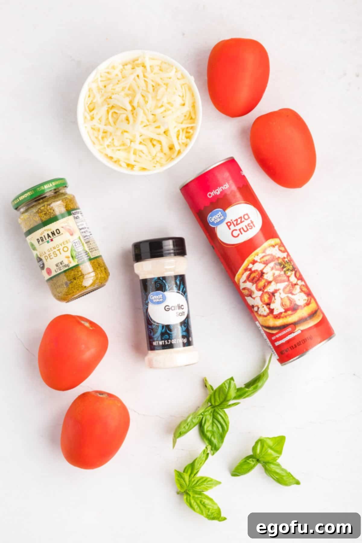 A flat lay photograph displaying all the fresh ingredients needed for Slab Tomato Pie: pizza dough, a jar of pesto sauce, sliced Roma tomatoes, shredded mozzarella cheese, a shaker of garlic salt, and a bunch of fresh basil leaves.