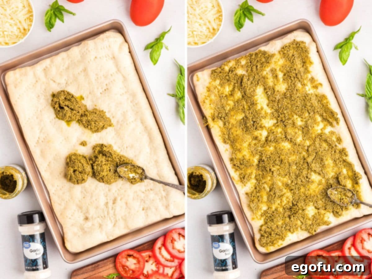 Hands spreading bright green pesto sauce evenly over a golden par-baked pizza dough crust on a baking sheet.