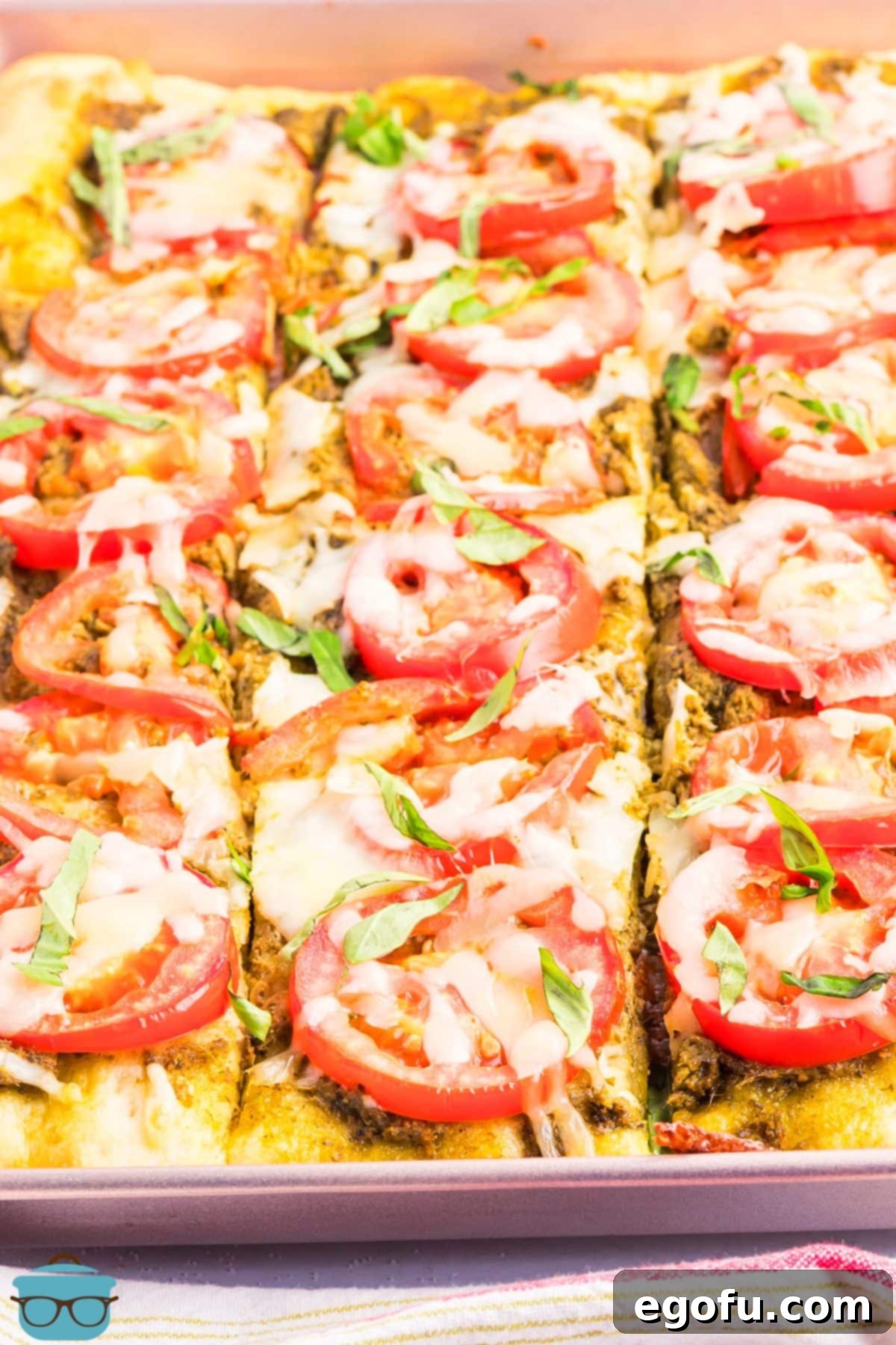 A close-up view of a freshly baked Slab Tomato Pie, already sliced into many square pieces, showing the crisp golden crust, melted cheese, roasted tomatoes, and fresh green basil on top.