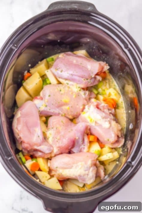 A Slow Cooker filled with chicken thighs, potatoes, carrots, celery, onion and minced garlic, with the broth mixture poured over it.