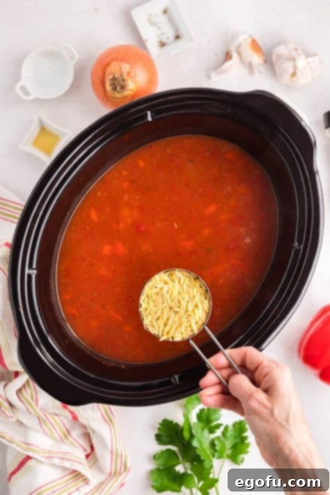 Uncooked orzo pasta being added to the simmering Chicken Parm Soup in the Crock Pot during the final cooking stage.