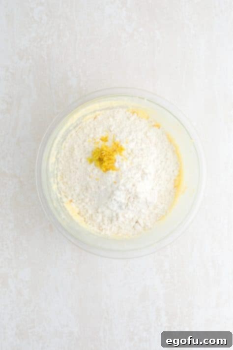 Flour and lemon zest being folded into the wet ingredients in a bowl.