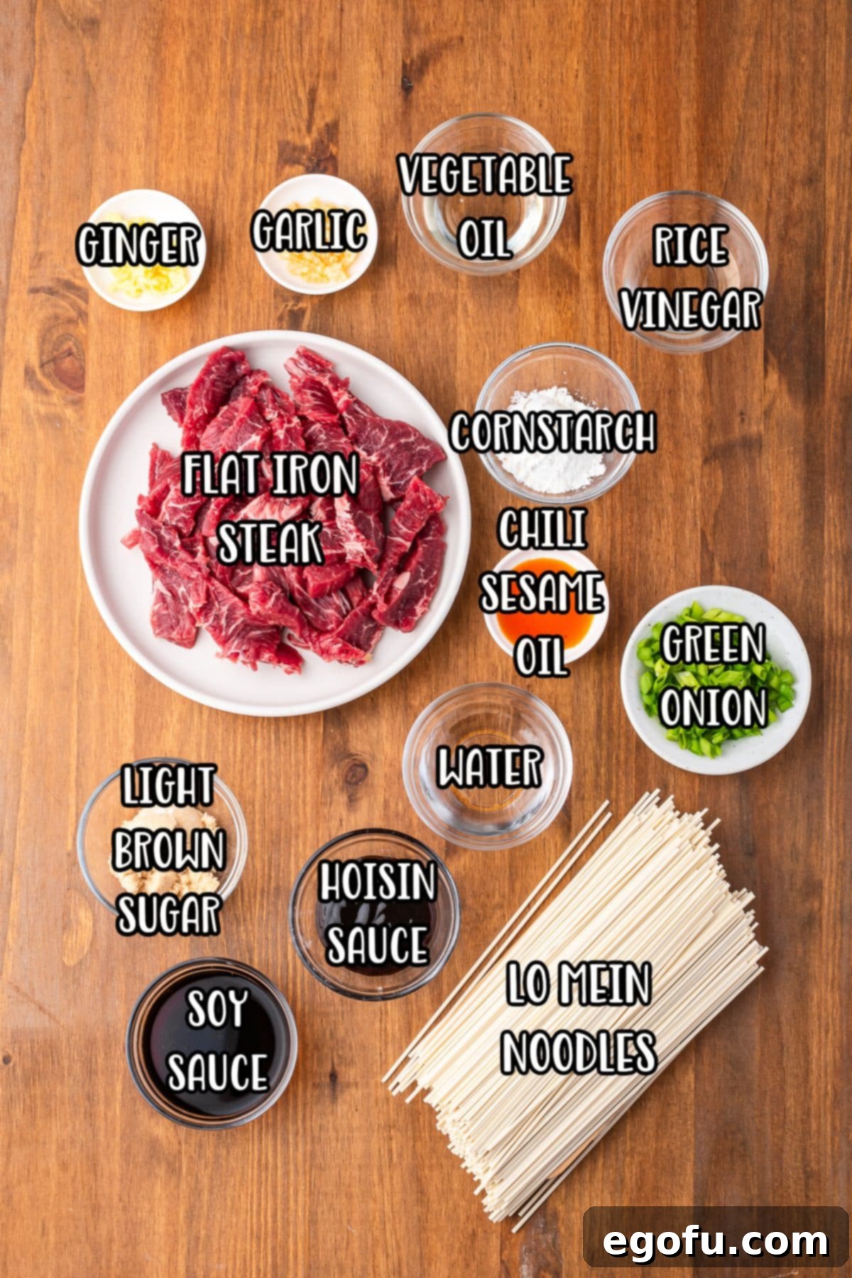 Arrangement of fresh ingredients for Mongolian Beef Noodles including ginger, minced garlic, sesame oil, rice vinegar, thin-sliced flat iron steak, cornstarch, chili sesame oil, green onion, water, hoisin sauce, light brown sugar, soy sauce, and lo mein noodles.
