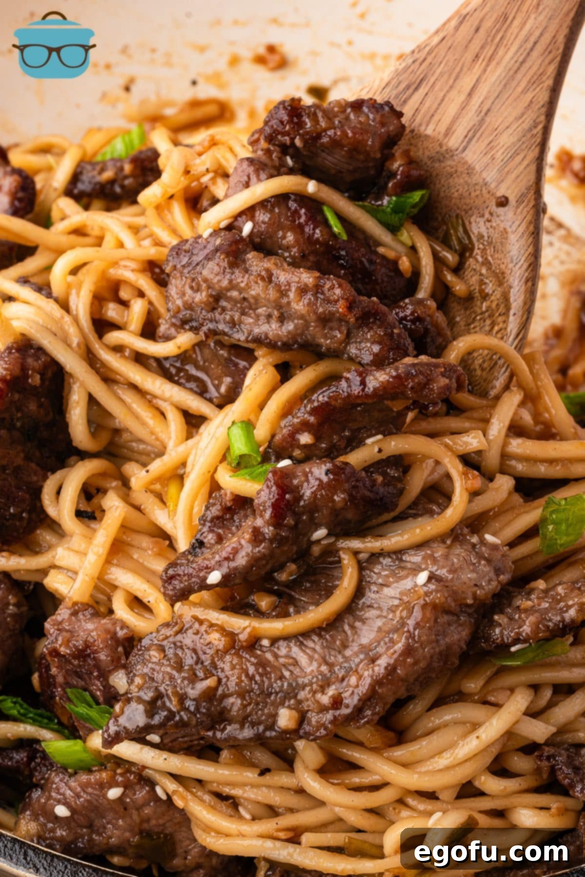 A close-up view of a wooden serving spoon stirring the Mongolian Beef Noodles in a large pot, showing the perfectly coated noodles and tender beef slices.