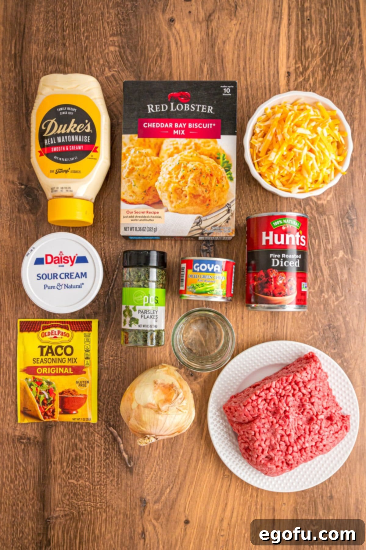 A flat lay image displaying all the ingredients needed for Cattle Drive Casserole, including ground beef, Red Lobster Cheddar Bay Biscuit Mix, water, onion, taco seasoning, fire roasted tomatoes, diced green chiles, shredded cheese, mayo, sour cream, and dried parsley.