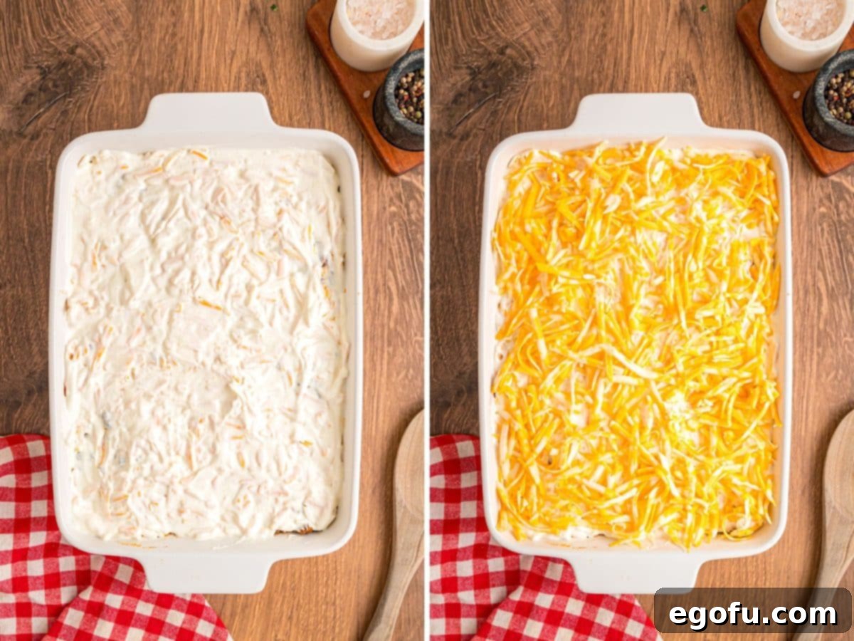 A layered casserole being assembled, with the creamy topping spread over the beef layer and then sprinkled with the final layer of shredded cheese.