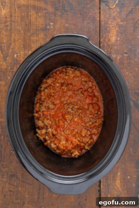 The initial layer of savory meat sauce carefully spread at the bottom of a slow cooker, forming the foundation for the baked ziti.