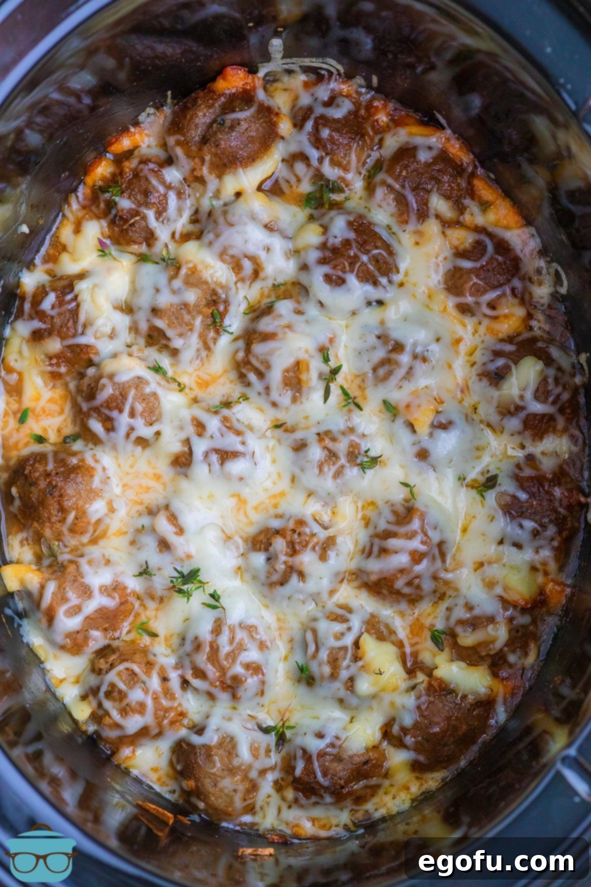 Looking down on a slow cooker filled with Crock Pot French Onion Meatballs, generously topped with melted, bubbly mozzarella cheese and a sprinkle of fresh herbs.